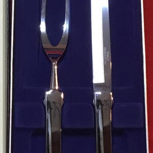 May include: A silver carving set with a knife and fork in a blue velvet box. The knife has a black handle and the text "TOSLER STAINLESS-STEEL" printed on the blade.