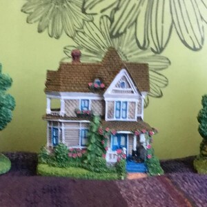 May include: Three miniature houses with painted details. The houses are blue, brown, and red with white trim. Each house has a small yard with trees and flowers.