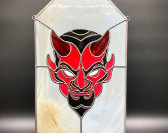 May include: A stained glass window depicting a red devil's face with black horns and eyes. The background is white with a slight iridescent sheen.