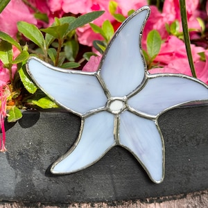 May include: A five-pointed starfish ornament crafted from pale lavender stained glass with a smooth, opalescent finish. The individual glass segments are joined together by thin, dark metallic solder lines, creating a clean, defined outline.