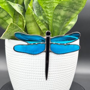 May include: A stained glass dragonfly with vibrant blue wings and a black body is displayed. The dragonfly is placed in a white textured pot with a green plant. The dragonfly is a decorative garden or home decor item.