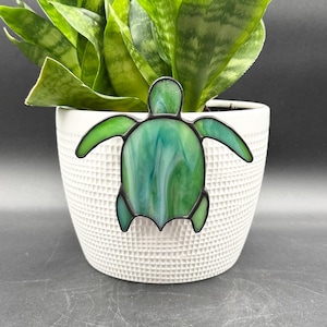 May include: A green and blue stained glass turtle decoration with a black outline is placed in front of a white textured pot with a green plant. The turtle has a marbled appearance and is a decorative accent.