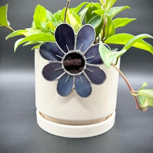 May include: A stained glass flower with dark blue and purple petals and a silver border. The flower is placed in a white ceramic pot with a plant inside. The plant has green leaves and is in front of a dark background.