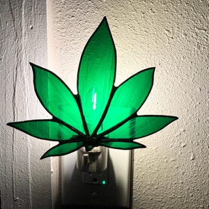 May include: A green glass cannabis leaf night light plugged into an electrical outlet. The light is on and glowing green.
