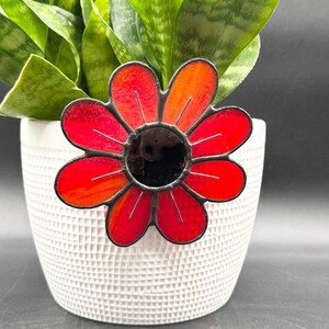May include: A stained glass flower with red and orange petals and a black center. The flower is placed in a white textured pot with a green plant in the background. The flower is a decorative garden accent.