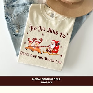 May include: A beige t-shirt with a Christmas-themed design featuring Santa Claus riding a sleigh pulled by reindeer. The text "Ho Ho Hold Up Gotta Take This Walkie Call" is printed in red on the shirt.