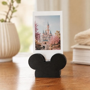 May include: A black wooden photo holder shaped like a mouse head silhouette, featuring a vertical slot that holds a rectangular photograph of a castle surrounded by pink flowering trees and a body of water under a soft, light-filled sky.