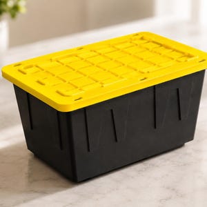 Puede incluir: Un contenedor de almacenamiento rectangular con una base negra sólida y una tapa de color amarillo brillante. La tapa presenta un patrón texturizado tipo rejilla con secciones rectangulares hundidas. El contenedor tiene un diseño cónico.