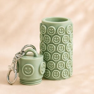 May include: A sage green cylindrical container and a matching cap, both featuring a repeating embossed smiley face pattern. The cap includes a silver-toned metal lobster clasp keychain attached to a small loop on its top surface.