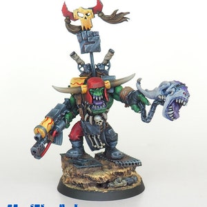 May include: A detailed miniature figure of a green-skinned orc warrior, holding a weapon and a grotesque head. The figure is painted with vibrant colors, including red, yellow, and blue accents. It stands on a textured base with a banner and skull details.