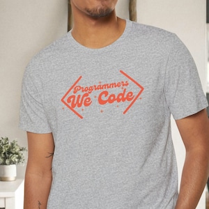 Puede incluir: Una camiseta gris con un gráfico rojo en forma de diamante que dice "Programmers We Code".