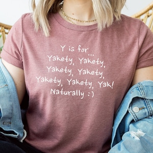 以下が含まれることがあります： 白文字で「Y is for... Yakety, Yakety, Yakety, Yakety, Yakety, Yakety, Yak! Naturally :)」と書かれた薄紫色のTシャツを着た女性。彼女は籐製の椅子に座り、トンボの模様が描かれた緑色のマグカップを持っている。