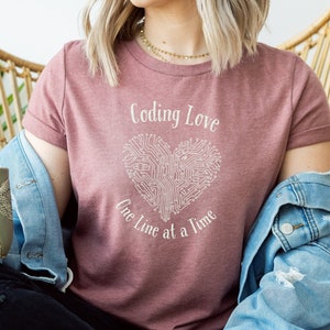 Könnte beinhalten: Eine Frau trägt ein mauvefarbenes T-Shirt mit dem Text "Coding Love One Line at a Time" und einer weißen Herzgrafik. Die Herzgrafik besteht aus Leiterplattenlinien. Sie sitzt auf einem Korbstuhl und hält eine grüne Tasse mit einem Libellenmuster.