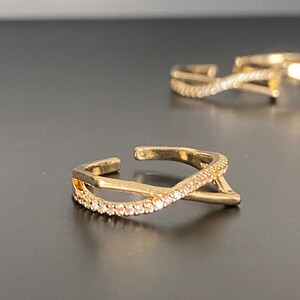 Può includere: Tre anelli color oro con un design a incrocio. Gli anelli sono aperti e adornati con piccole pietre scintillanti. Gli anelli sono esposti su una superficie scura.