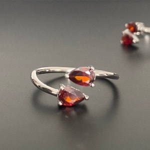 May include: A silver ring with two teardrop-shaped red gemstones. The ring is open and adjustable.