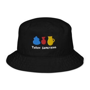 以下が含まれることがあります： 青、赤、黄色の陶器のデザインが刺繍された黒いバケットハット。「Throw Something」の文字が白で刺繍されています。つばが広く、柔らかい素材で作られています。