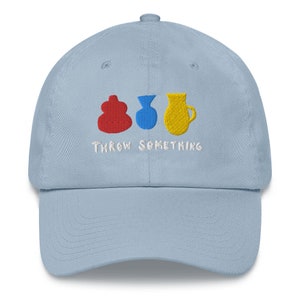 以下が含まれることがあります： 刺繍が施された水色の野球帽。赤、青、黄色の3つのカラフルな陶器の形がデザインされており、下に「THROW SOMETHING」の文字が刺繍されています。帽子はカーブしたつばと調節可能なストラップが付いています。