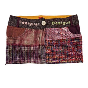 以下が含まれることがあります： ライトピンクとグリーンの刺繍で「Desigual」の文字が入ったダークブラウンのウエストバンドが特徴のパッチワークミニスカート。メタリックカッパーの生地、パープルのチェック柄ツイード、マルチカラーの織物など、さまざまな質感を組み合わせています。