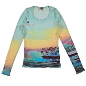 May include: A long-sleeved, light blue shirt with a watercolor-like print of a sunset and ocean scene. The shirt has the text "The Month Total Solar Cop... Copine Paris" printed on the front.