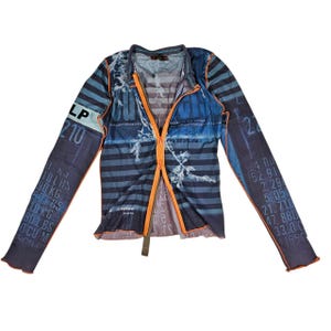 Puede incluir: Top azul y naranja de manga larga con cremallera y estampado de rayas. Las mangas presentan texto y números. El top tiene cuello en V y cremallera naranja en contraste. El diseño general es una mezcla de tonos azules con detalles blancos.