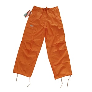 Könnte beinhalten: Leuchtend orangefarbene Cargohose mit mehreren Taschen und Kordelzügen an Taille und Knöcheln. Ein kleines Etikett mit dem Wort "UFO" und einem Flaggenmuster ist angebracht. Die Hose hat eine lockere Passform.