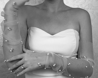 May include: A pair of sheer, white bridal gloves with pearl embellishments. The gloves are worn on the arms of a person wearing a strapless white dress.
