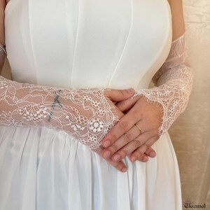 May include: A close-up of a white wedding dress with long, delicate lace sleeves. The sleeves are intricately patterned with floral designs. A gold ring is visible on the ring finger. The dress is made of a smooth, white fabric.