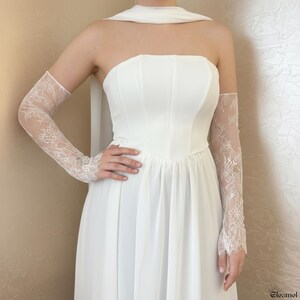 May include: A white strapless wedding dress with a flowing skirt and matching sheer lace gloves. The gloves extend from the wrist to the upper arm, featuring intricate floral lace patterns. The dress has a fitted bodice.