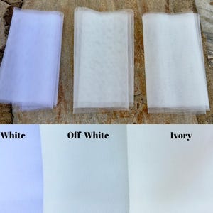 May include: Three swatches of tulle fabric in white, off-white, and ivory. The swatches are laid out on a textured surface.