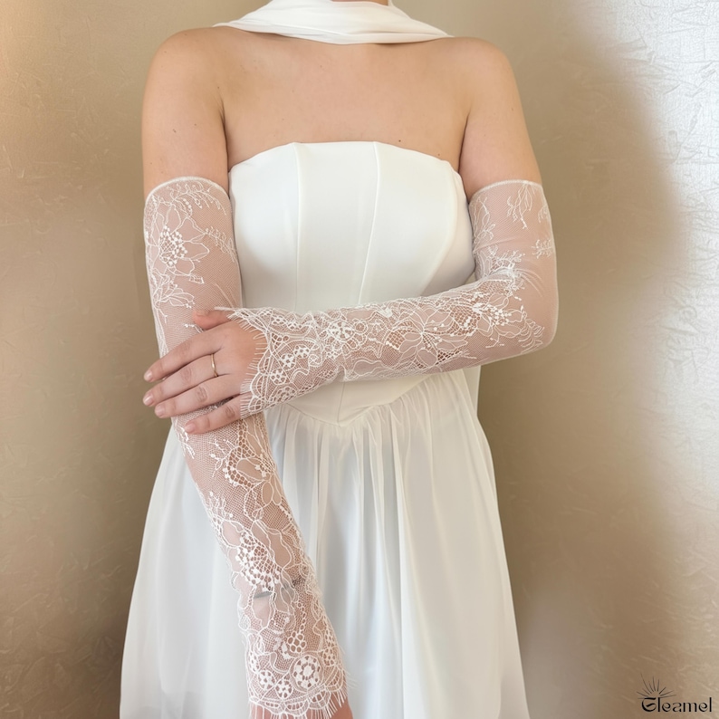 May include: A white strapless dress with matching lace arm gloves. The gloves are long, covering the arms and hands, and feature a delicate floral lace pattern. The dress has a fitted bodice and a flowing skirt.