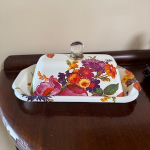 May include: A white butter dish with a floral design featuring purple, orange, and yellow flowers. The dish has a clear glass knob on top and a matching tray. The butter dish is on a dark wooden surface.