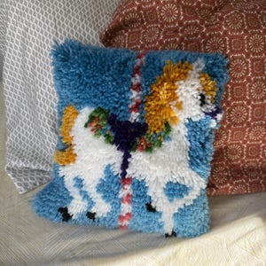 May include: A square, blue, handmade pillow with a white unicorn design. The unicorn has a yellow mane and tail, with a purple and green floral garland. The pillow is made of a textured, tufted material.