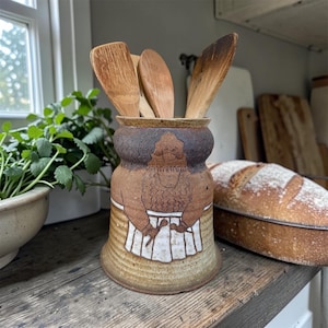 May include: A ceramic utensil holder with a brown and tan color scheme. The holder features a hand-drawn design of a person wearing an apron. Wooden spoons and spatulas are inside the holder. The holder is on a wooden surface.