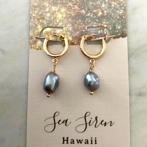 May include: Gold-tone hoop earrings with gray, teardrop-shaped pearls. The earrings hang from a card with the text "Sea Siren Hawaii" in elegant script. The background has a glittery, gold effect.