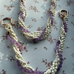 以下が含まれることがあります： 白い貝殻ビーズと紫色のビーズのストランドが特徴の、2つの手作りのハンドバッグストラップ。各ストラップには、取り付け用の金色の金属製Dリングが付いています。ストラップは、花柄の背景に表示されています。