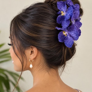 May include: A hair accessory featuring vibrant deep blue Vanda orchids with speckled petals and yellow centers, secured in a dark brown updo hairstyle. The accessory is paired with a gold hoop earring featuring a single white teardrop pearl.