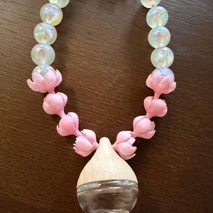 May include: A necklace featuring iridescent, clear beads and pink flower-shaped accents. The necklace has a clear glass vial with a wooden cap at the bottom and a silver clasp at the top. The necklace is designed to hold essential oils.