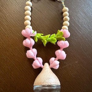 May include: A clear glass bottle with a wooden top, adorned with a pink floral garland and wooden beads. The bottle is attached to a gold-colored clasp. The floral garland has green leaves.