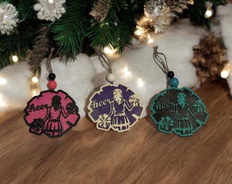 May include: Three wooden Christmas ornaments shaped like a cheerleader holding pom-poms. Each ornament is painted a different color: pink, purple, and teal. The word "cheer" is written on each ornament.