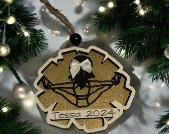 May include: Gold glitter Christmas ornament with a black silhouette of a gymnast doing a handstand on a bar. The ornament has a white bow and the text "Tessa 2024" on it.