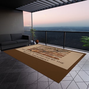 May include: A rectangular brown outdoor rug with a tan center featuring a rifle design and the text "THIS HOUSE IS PROTECTED BY THE SECOND AMENDMENT." The rug is on a tiled patio with a view of a cityscape.
