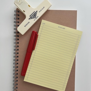May include: A brown spiral-bound notebook, a cream-colored plastic Penco Clampy clip, a red pen, and a yellow to-do list notepad with lines and checkboxes. The clip features the text Penco Clampy, PLA-CLIP, and BINDING. The notepad includes the text TO DO LIST.