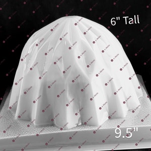 May include: White plastic mold for making a cake or dessert. The mold is 6 inches tall and 9.5 inches wide. It has a rounded top and a flat base.