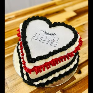 May include: A heart-shaped cake with white frosting, black and red decorative borders, and a calendar for August. The cake sits on a silver cake board and is displayed on a wooden surface.