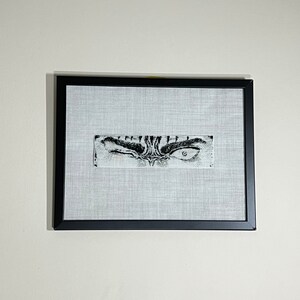 May include: A framed artwork with a black frame, showcasing a detailed black and white drawing of an eye. The artwork is rectangular, set against a light grey fabric. The eye is the focal point, with intricate details.