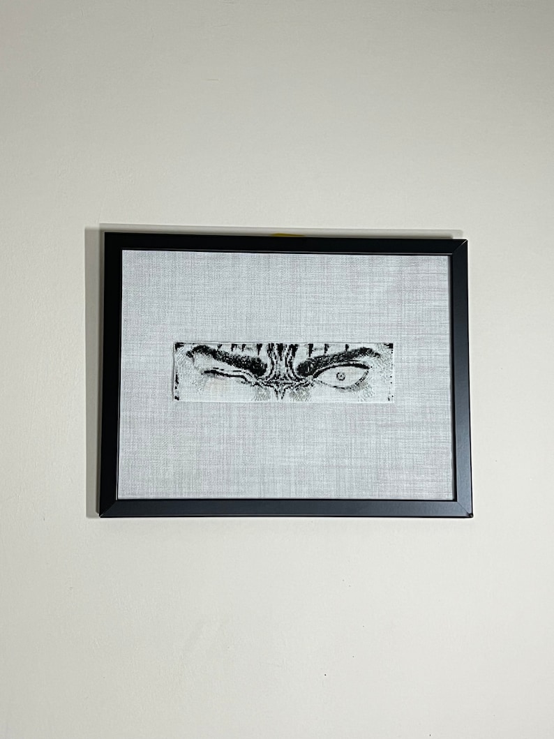 May include: A framed artwork with a black frame, showcasing a detailed black and white drawing of an eye. The artwork is rectangular, set against a light grey fabric. The eye is the focal point, with intricate details.