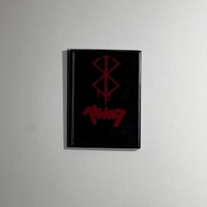 May include: A framed black artwork with a red symbol and text. The symbol is a geometric design with dripping elements. Below the symbol is red text. The artwork is mounted on a white wall.