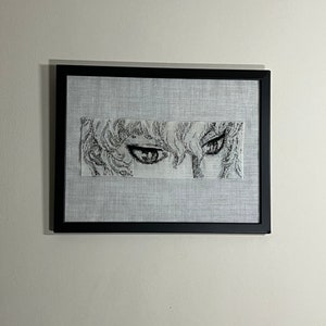 May include: A framed artwork showing a detailed black and white illustration of a character's eyes and hair. The artwork is set against a light grey fabric background within a black frame, creating a striking contrast. The piece is mounted on a white wall.