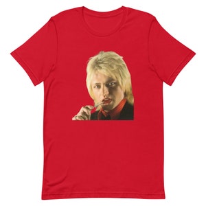 May include: A red t-shirt featuring a portrait of a person with blonde hair, wearing a black suit jacket and a red collared shirt, holding a lollipop. The image is centered on the shirt.