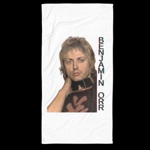 May include: A white beach towel with a photo of a man with long blonde hair and a pink and black shirt. The text "BENJAMIN ORR" is printed vertically in red on the right side of the towel.
